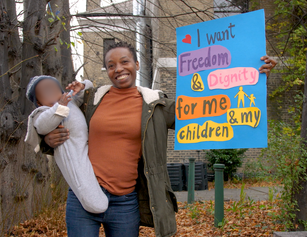 A woman holding a poster that says 'I want dignity and freedom for me and my children'. In her other hand, she is holding her baby - both are smiling.
